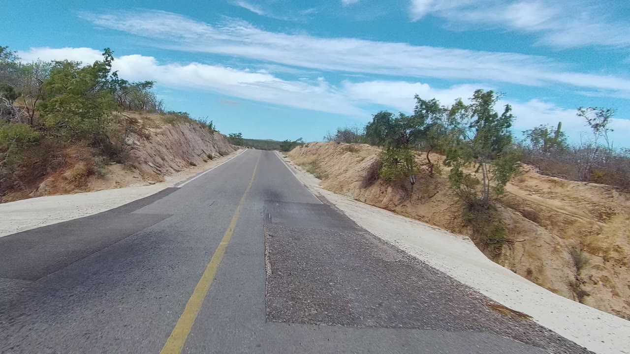 A paved East Cape Los Cabos road runs between dry cut slopes and sparse trees under scattered clouds, with no visible people.