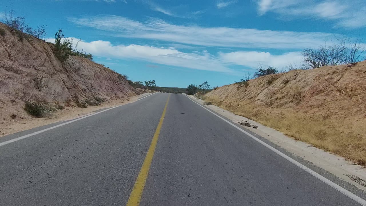 An empty asphalt road in East Cape Los Cabos passes eroded tan and gray hillsides with dry brush and no visible people.