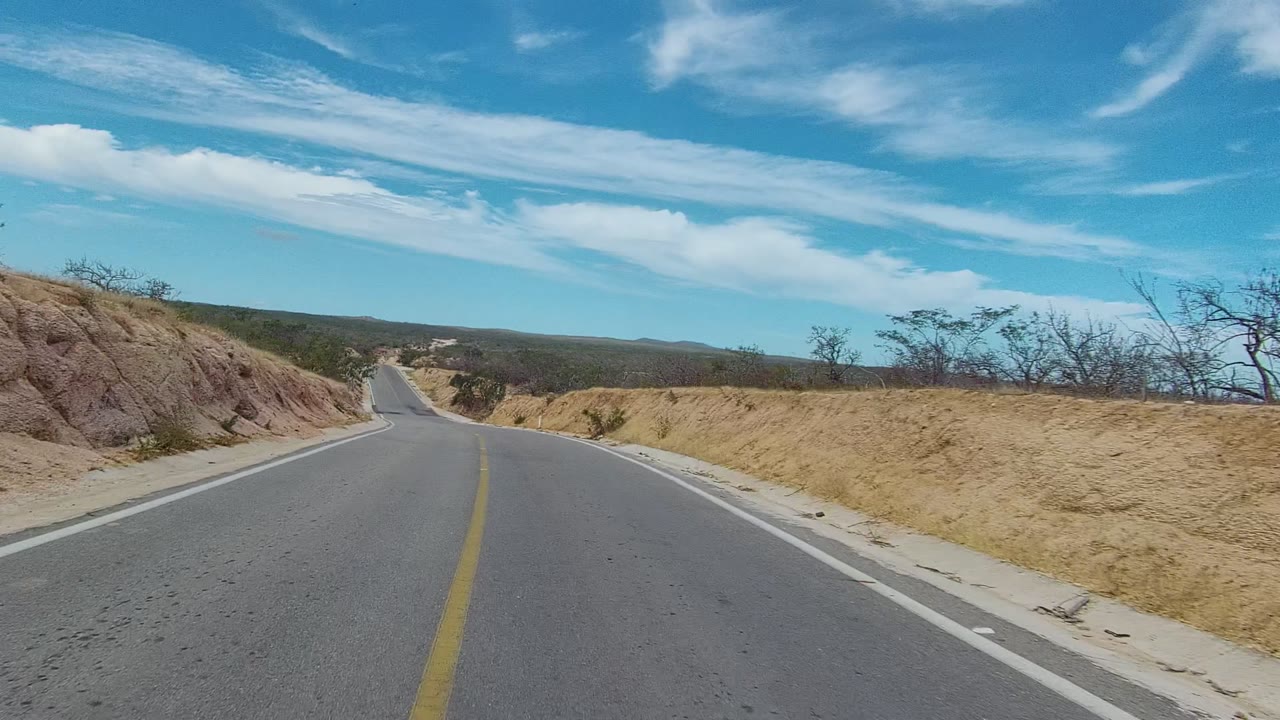 A straight paved road in East Cape Los Cabos descends between pale dry hillsides and sparse trees, with open hills in the distance and no visible people.