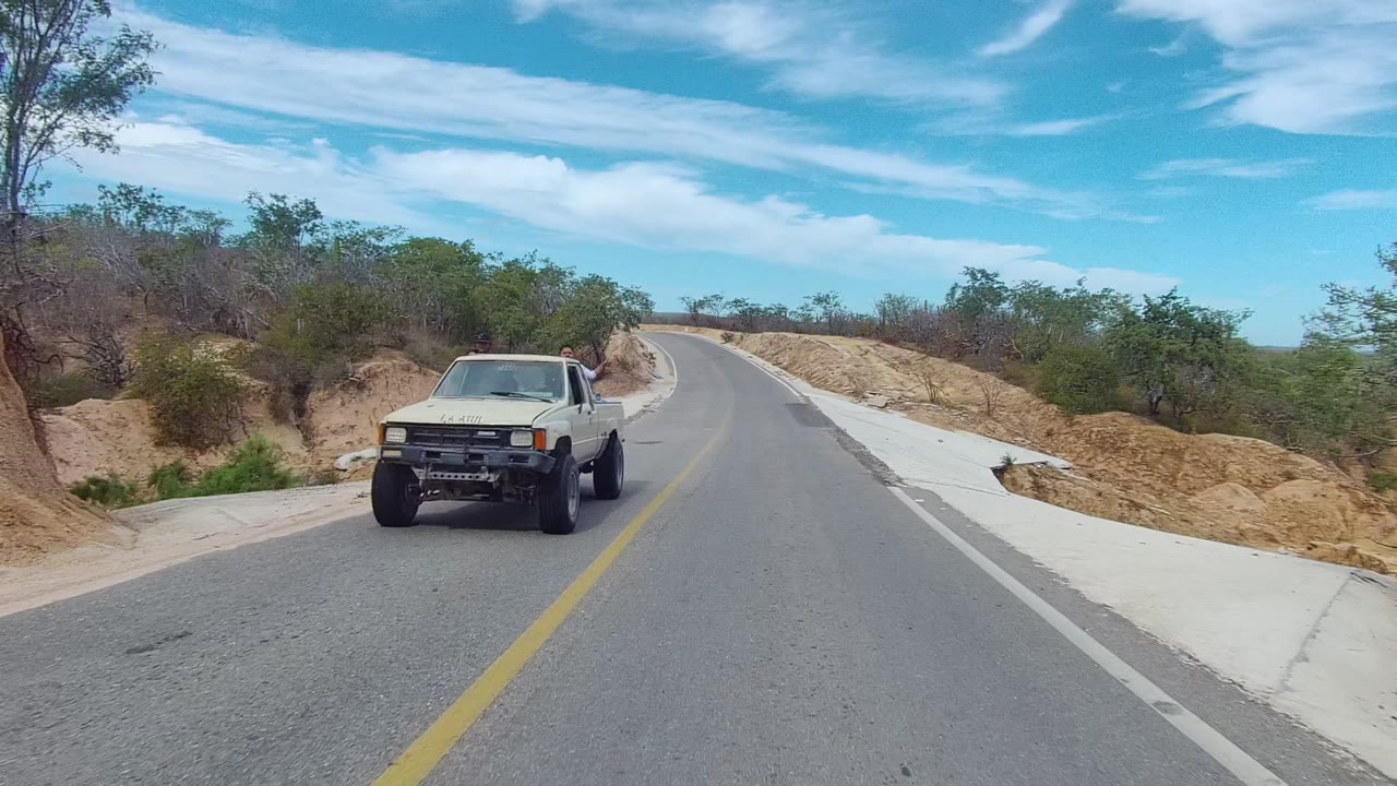 White pickup truck drives toward the left side of a rural road in East Cape Los Cabos, with dry brush, sandy banks, blue sky, and no visible people.