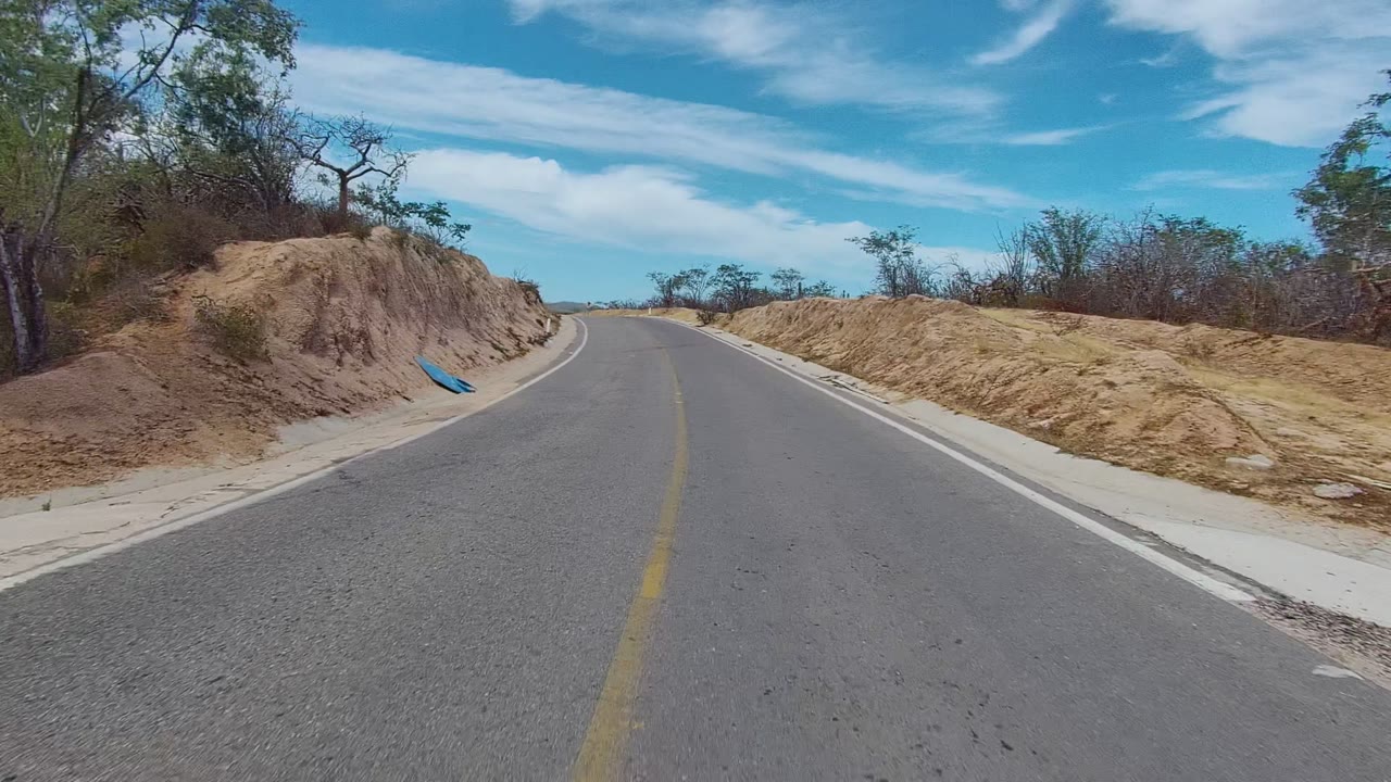 An empty East Cape Los Cabos road bends right between pale dirt embankments, scrubby trees, and a blue sky streaked with clouds, with no visible people.
