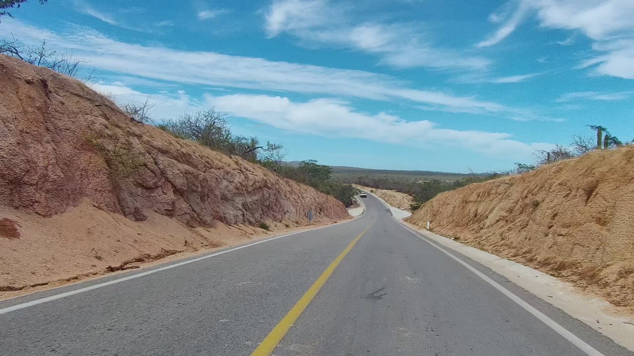 A road-level view in East Cape Los Cabos passes a tall reddish eroded bank on the left and a tan slope on the right beneath wispy clouds, with no visible people.