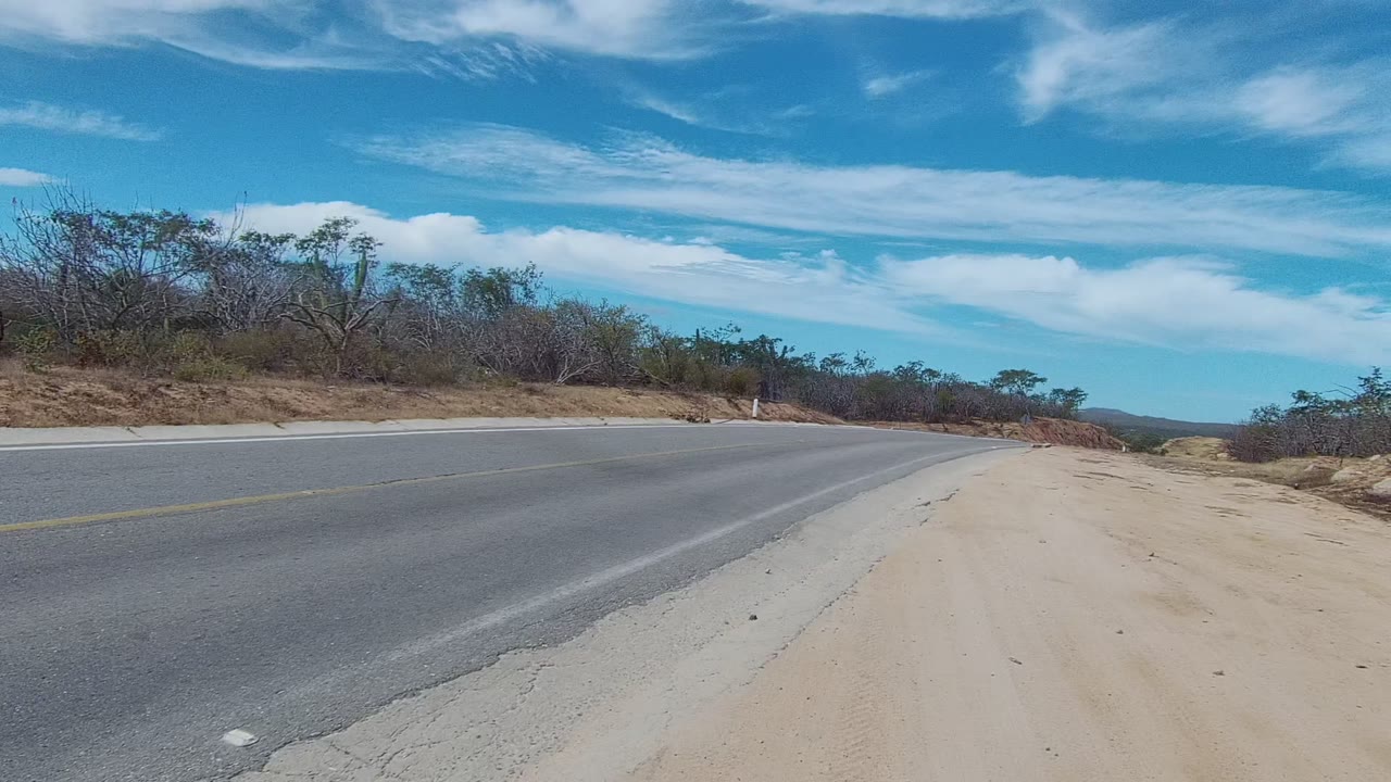 Paved road and pale dirt shoulder in East Cape Los Cabos, with scrubby roadside vegetation, scattered clouds, and no visible people.