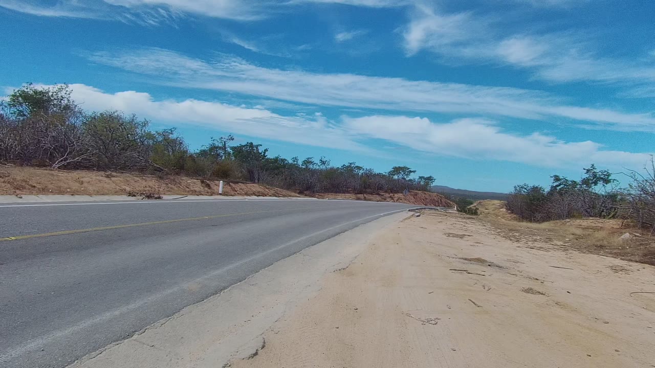 Empty paved road in East Cape Los Cabos with a sandy right shoulder, roadside scrub, distant low terrain, streaked white clouds, and no visible people.
