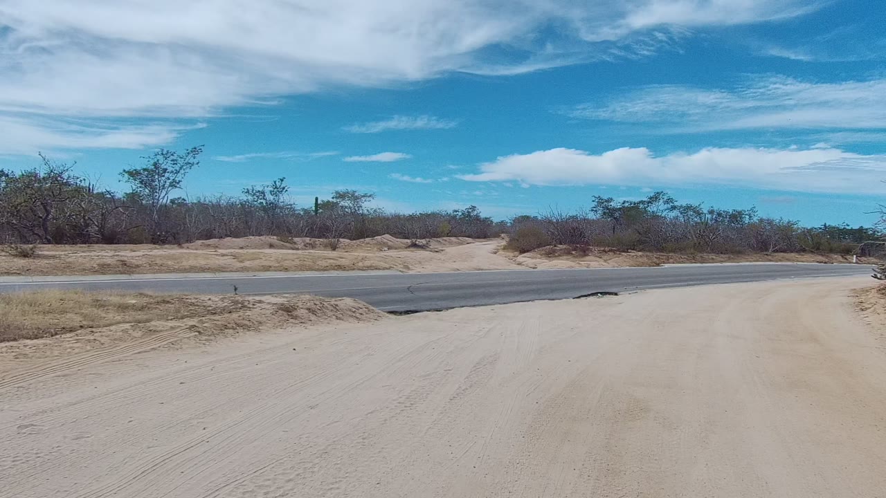 The sandy track nears a gray paved highway in East Cape Los Cabos, with scrub-covered berms, open sky, and no visible people.