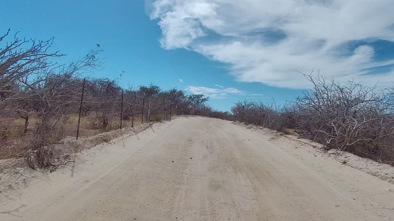 Opening view of a sandy dirt road in East Cape Los Cabos, Baja California Sur, with dry bushes on both sides, fence posts at left, white clouds overhead, and no visible people.