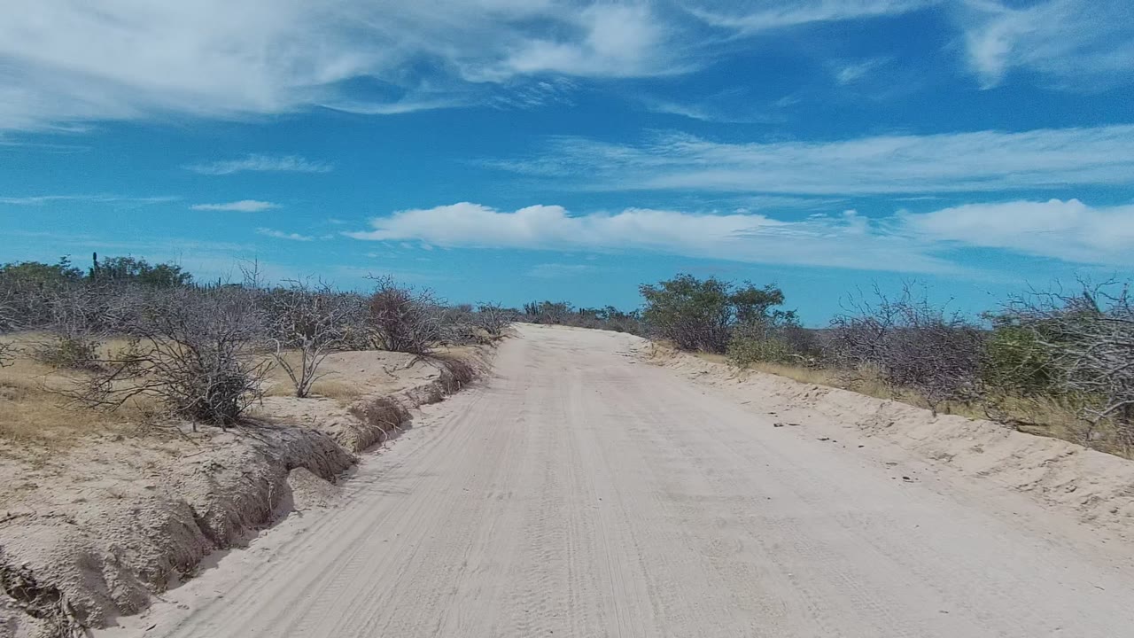 Sandy dirt road in East Cape Los Cabos, Baja California Sur, with low dry bushes on both sides, scattered clouds overhead, and no visible people.