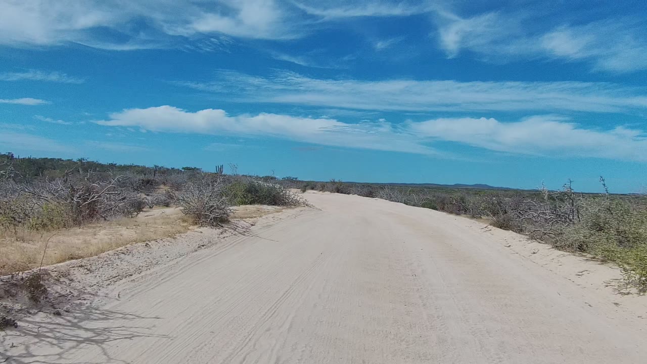 A wide sandy track in East Cape Los Cabos runs toward low distant terrain, with scrub and dry brush on both sides and no visible people.
