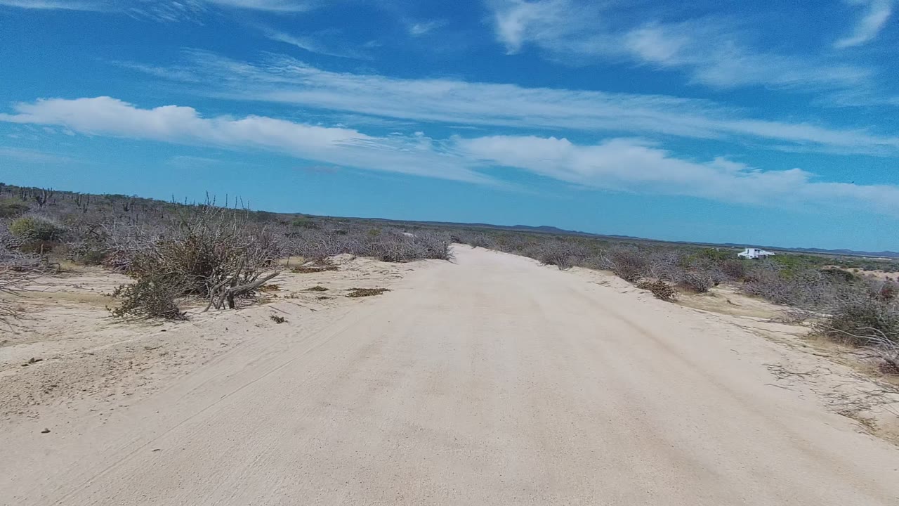 A sandy road in East Cape Los Cabos runs straight ahead between dry bushes and low scrub under a blue sky with scattered clouds, with no visible people.