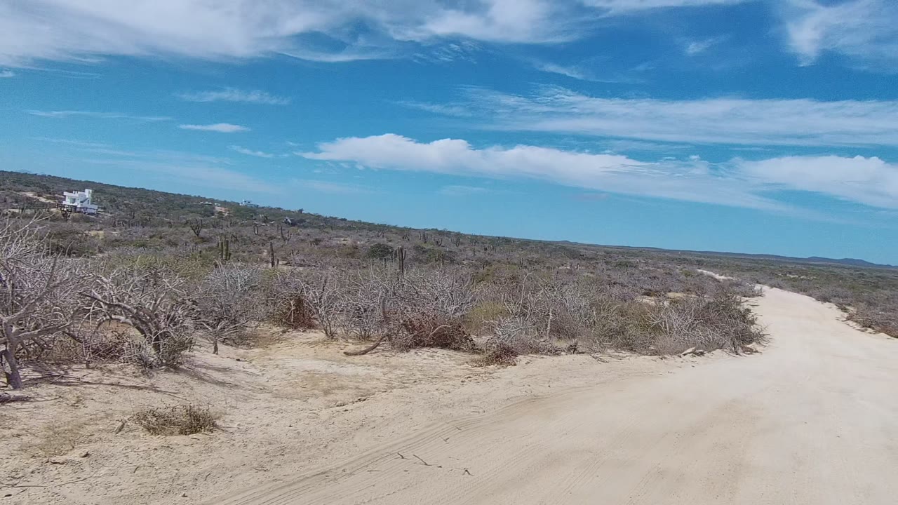 At the start of the clip, a sandy dirt road passes through arid East Cape Los Cabos scrubland with dry bushes, cacti, distant low hills, and no visible people.