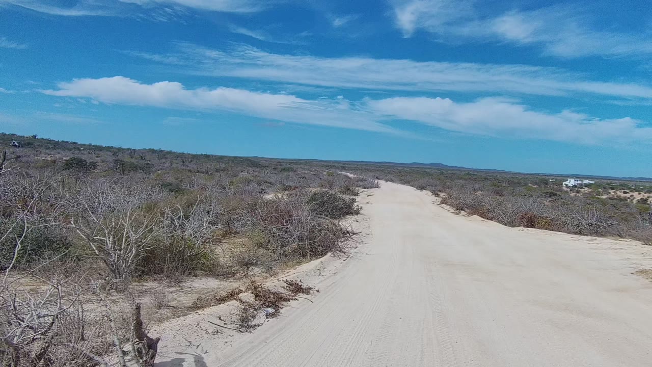 At 0:02, the wide sandy road curves through scrubland, with tangled dry brush in front, low hills on the horizon, a small white structure in the distance, and no visible people.