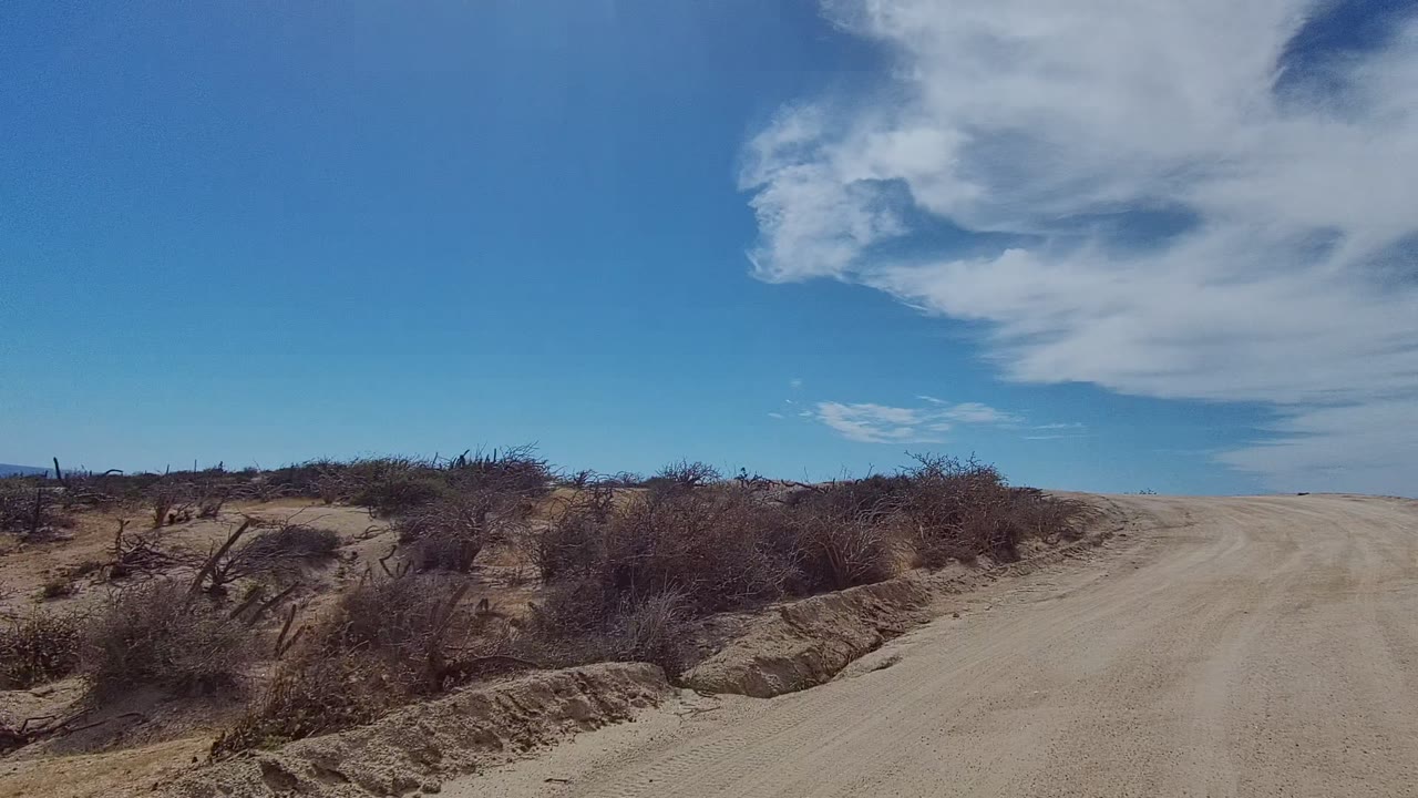 Sandy dirt road in East Cape Los Cabos, Baja California Sur, Mexico, bordered by dry bushes and eroded sandy shoulders under broad white clouds, with no visible people.