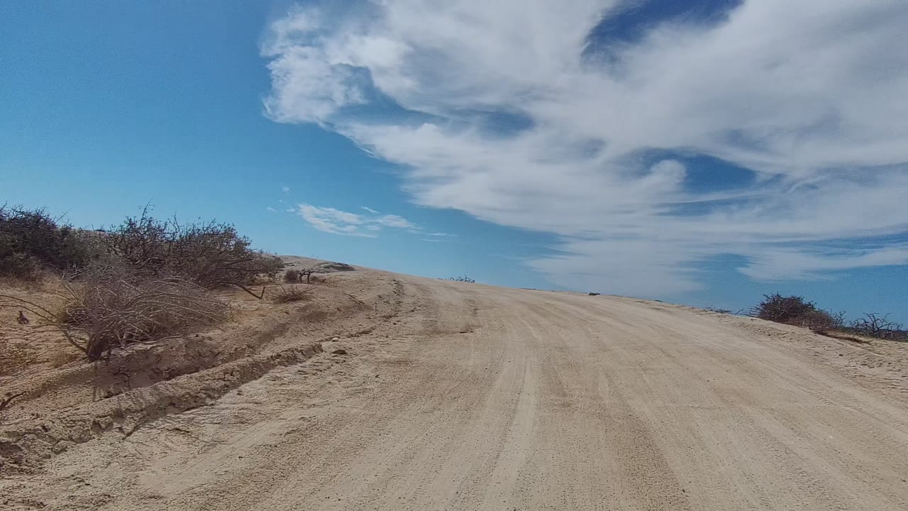 The sandy road fills the foreground in East Cape Los Cabos as it approaches a low crest, with dry shrubs on the left and right and large white clouds across the blue sky, no visible people.
