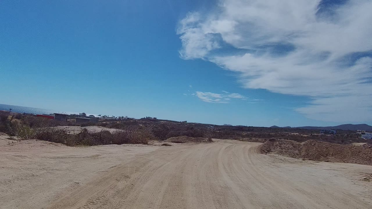Wide view of a sandy dirt road through arid East Cape Los Cabos scrubland with distant hills, scattered clouds, and no visible people.