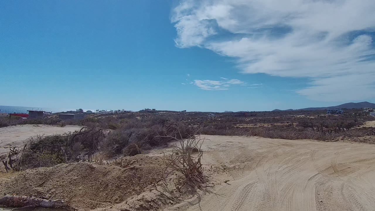 Sandy track crossing dry East Cape Los Cabos terrain with brush piles close to the road, low hills beyond, scattered buildings, and no visible people.