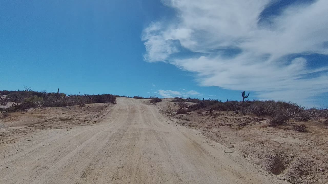 At 0:00, a sandy dirt road in East Cape Los Cabos, Baja California Sur rises through low desert brush with cacti on the horizon and no visible people.