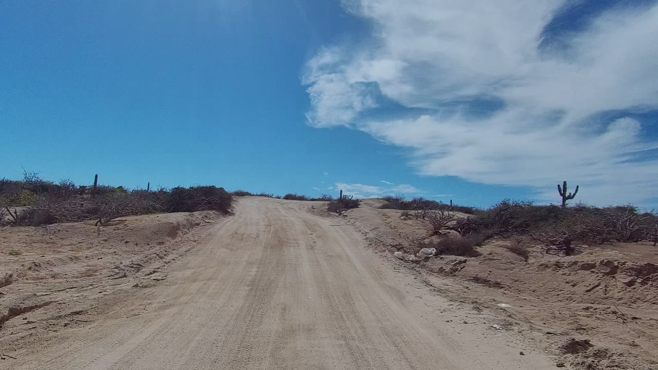 Dirt road continues uphill with scrub on both sides and clouds overhead.