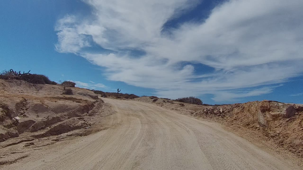 East Cape Los Cabos dirt road begins beside eroded sandy banks and sparse desert brush under a broad blue sky, with no visible people.