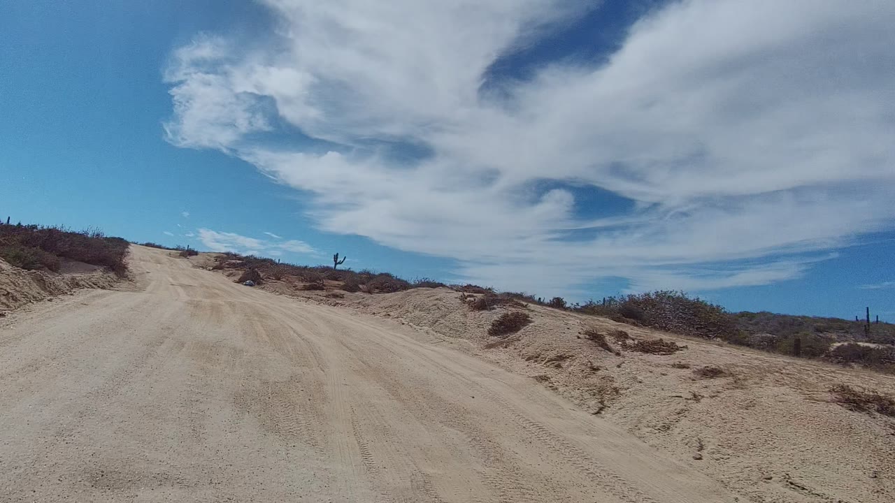 East Cape Los Cabos desert road fills the foreground with rutted sand, scrub-covered banks, scattered cacti near the horizon, and no visible people.