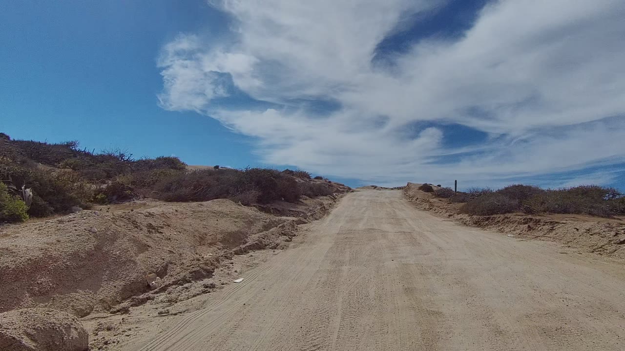 A centered sandy dirt road in East Cape Los Cabos, Mexico, rises between scrubby banks under broad white clouds, with no visible people.