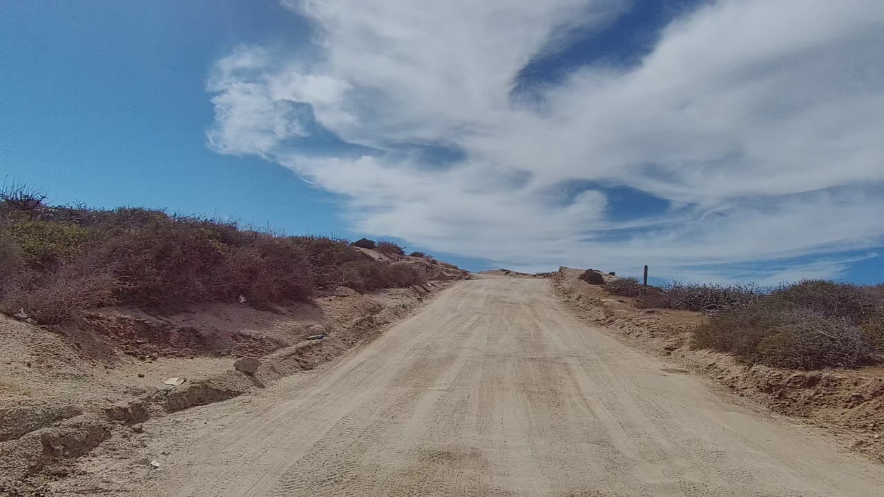 Dusty uphill road bordered by scrub and sandy cut banks beneath cloud cover.
