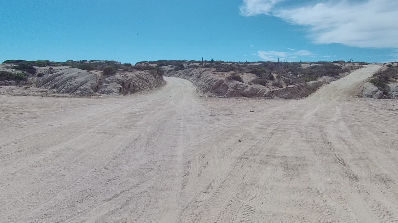 Wide opening view of a sandy dirt road in East Cape Los Cabos, Baja California Sur, with low eroded dunes, sparse scrub, tire tracks, scattered clouds, and no visible people.