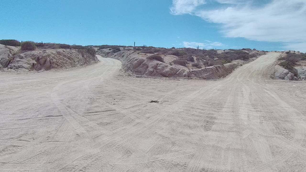 Dirt road curves between pale sandy mounds with scrub on the ridge line.