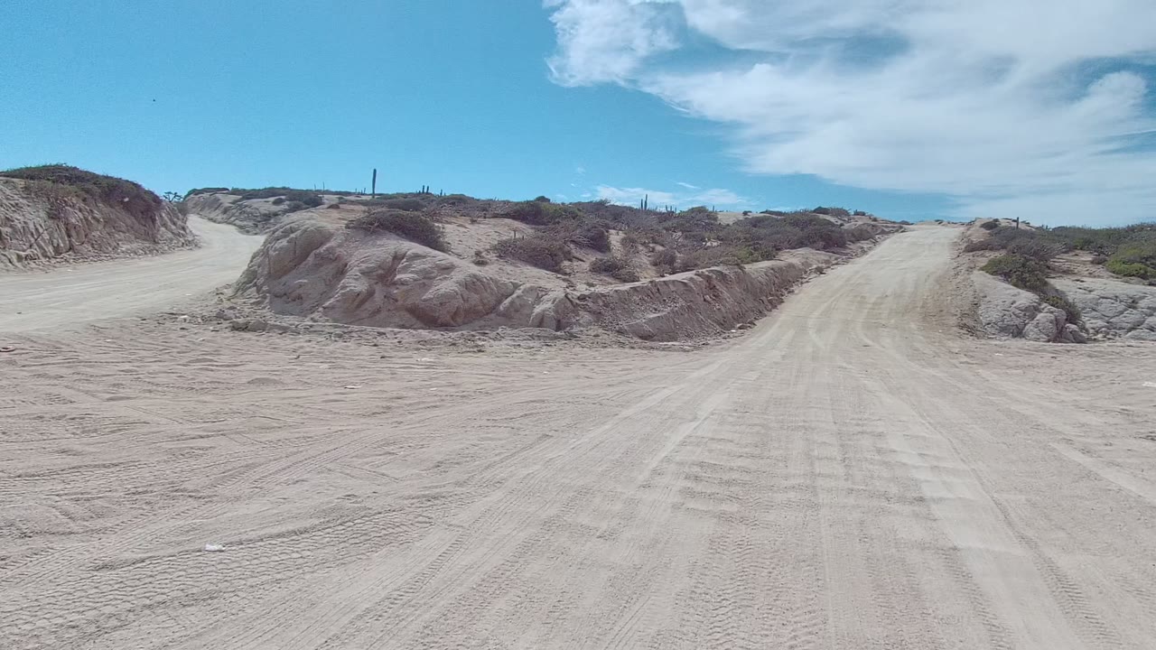 Midwide frame from East Cape Los Cabos, Baja California Sur, with the sandy road crossing the foreground, low dune ridges on both sides, scattered scrub, a bright blue sky, and no visible people.