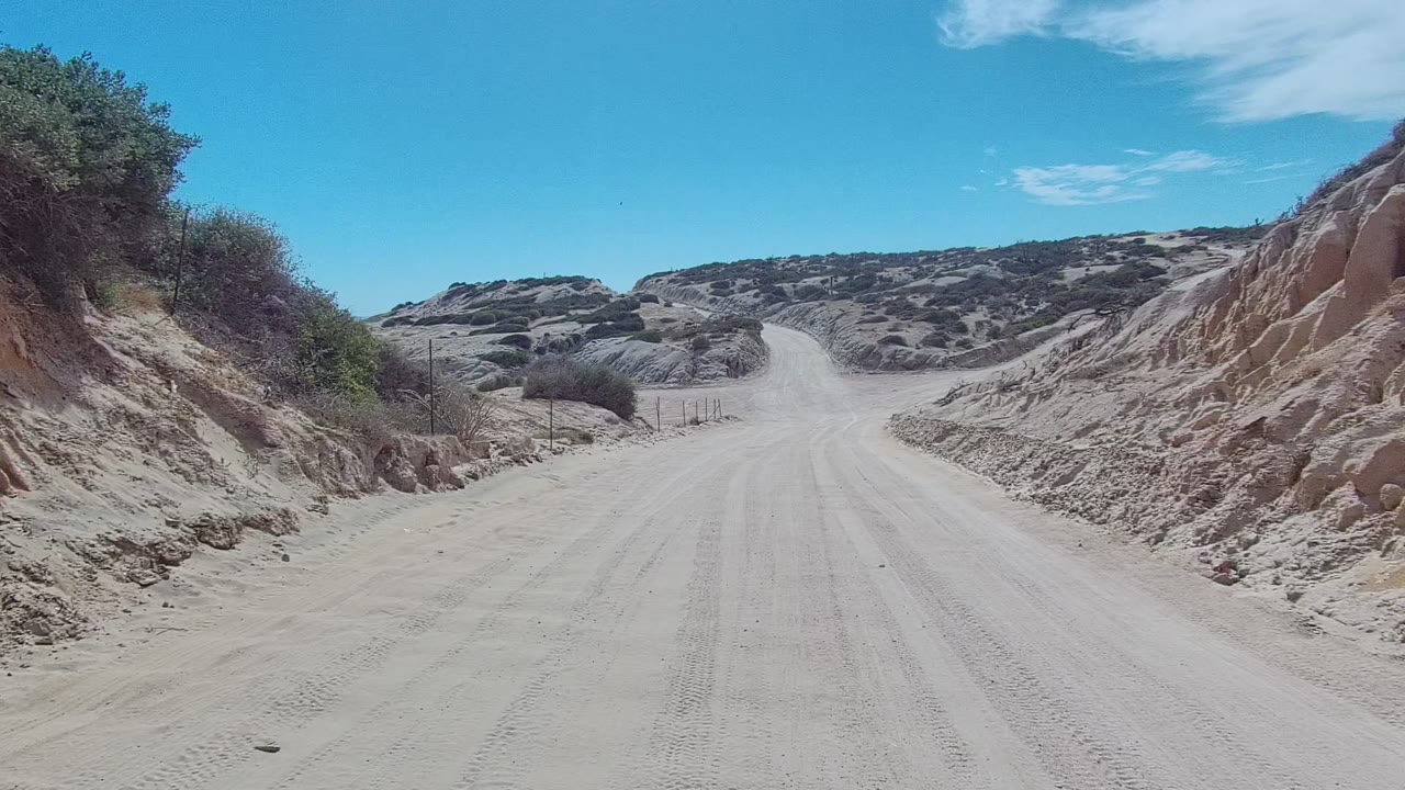 Start of the East Cape Los Cabos clip showing a pale dirt road between sandy banks, scrub vegetation, and blue sky, with no visible people.
