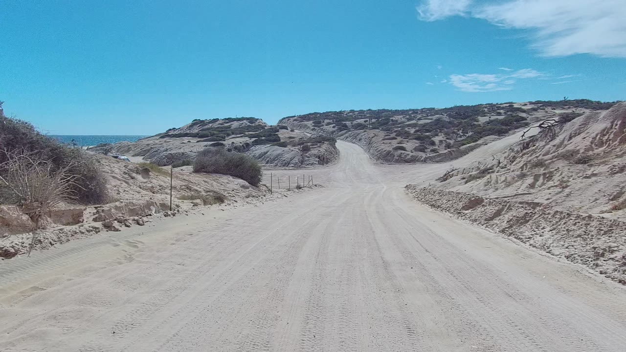 Sandy road curving through low desert hills with a small ocean glimpse.