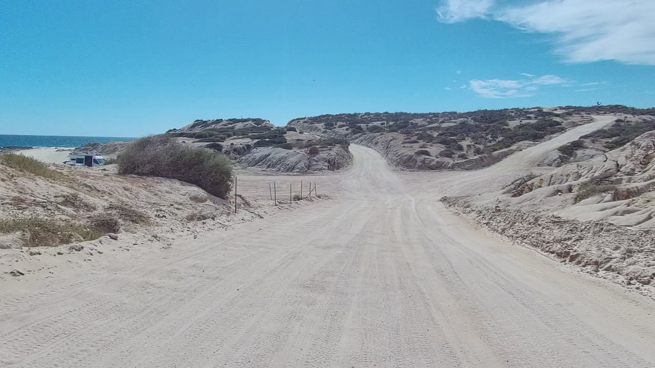 The road opens wider in East Cape Los Cabos, with fence posts, pale sand, scrub-covered hills, a blue sky, and no visible people.