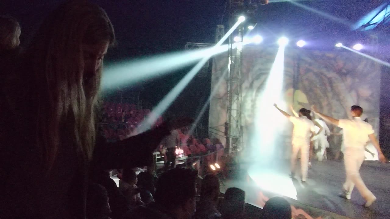 In San Jose del Cabo, dancers in white perform on stage under bright white spotlights while audience members appear as dark foreground silhouettes.