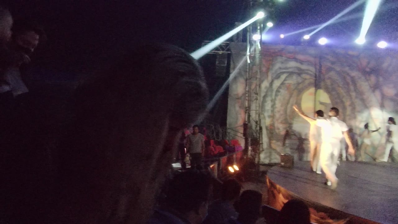 White-clad performers dance under bright beams with audience in shadow.