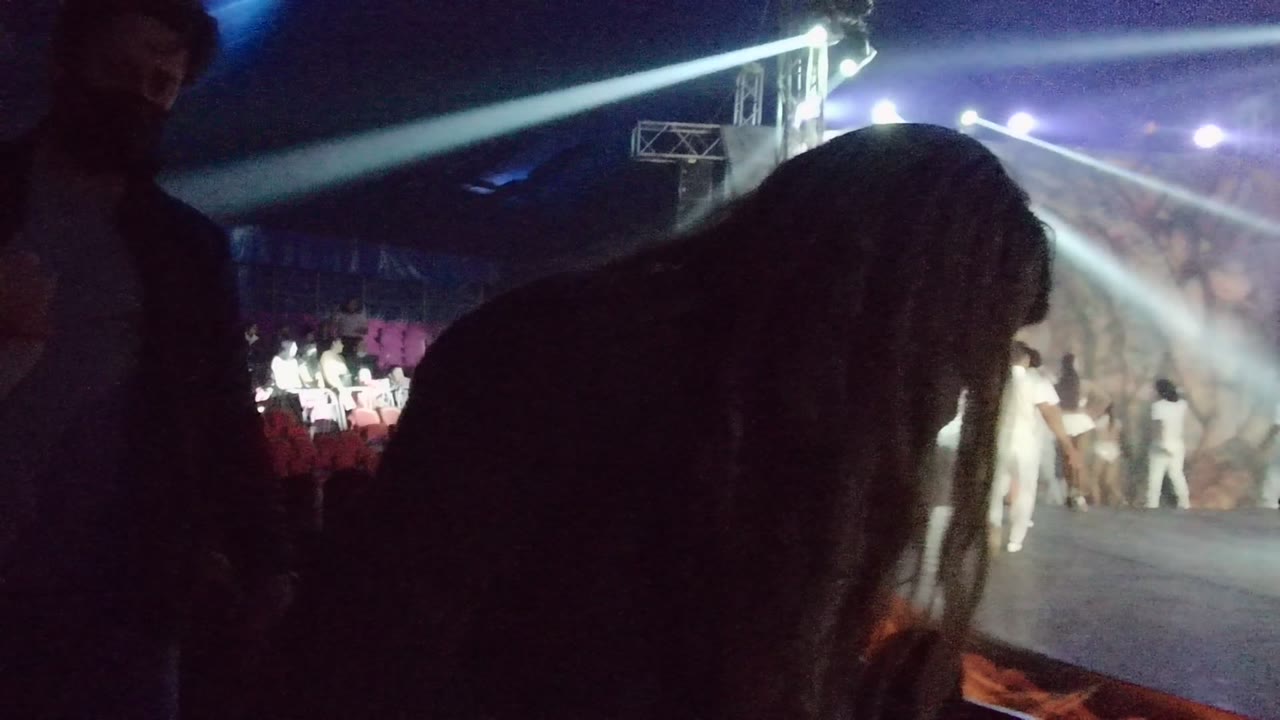 In San Jose del Cabo, a large audience silhouette partially blocks the stage while dancers in white continue performing beneath white spotlights.