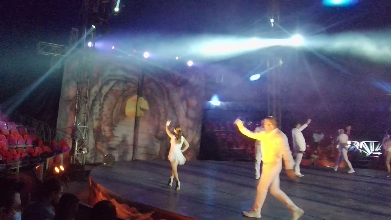 In San Jose del Cabo, dancers in white costumes perform on a stage with audience silhouettes in front, red seating behind them, and bright beams crossing the stage.