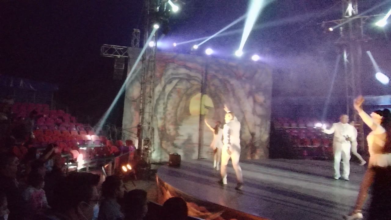 Multiple dancers in white spread across the San Jose del Cabo stage as a female performer enters at the right edge and purple haze surrounds the spotlights.