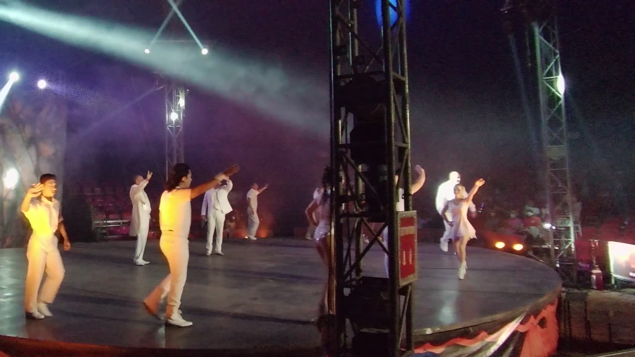 Dancers in white outfits perform on a dark stage in San Jose Del Cabo, Mexico, with a side-stage truss in the foreground and blue-purple spotlights above.