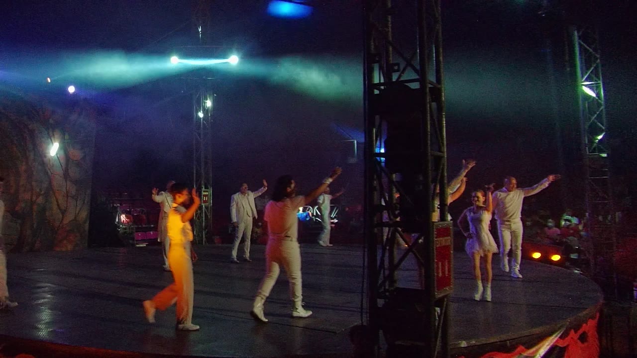 White-clad performers gesture on stage beside a central lighting truss.