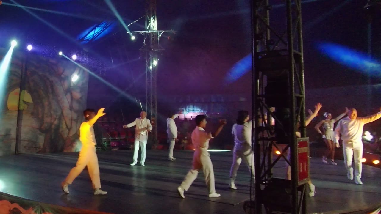A group of dancers in white perform under green-blue beams on the San Jose Del Cabo stage, with warm lights low on the floor and a scenic backdrop behind them.
