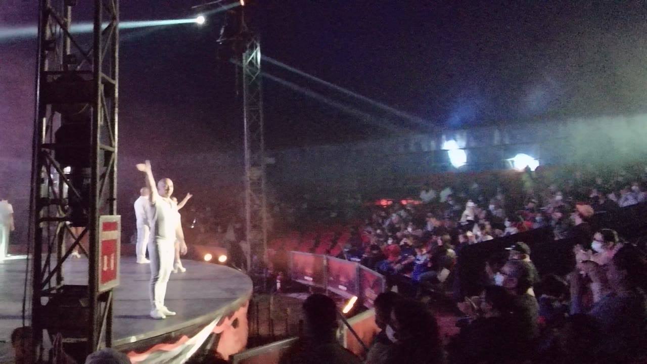 White-costumed dancers perform on a round stage in San Jose Del Cabo, Mexico, with audience members in the foreground and colored haze above the crowd.