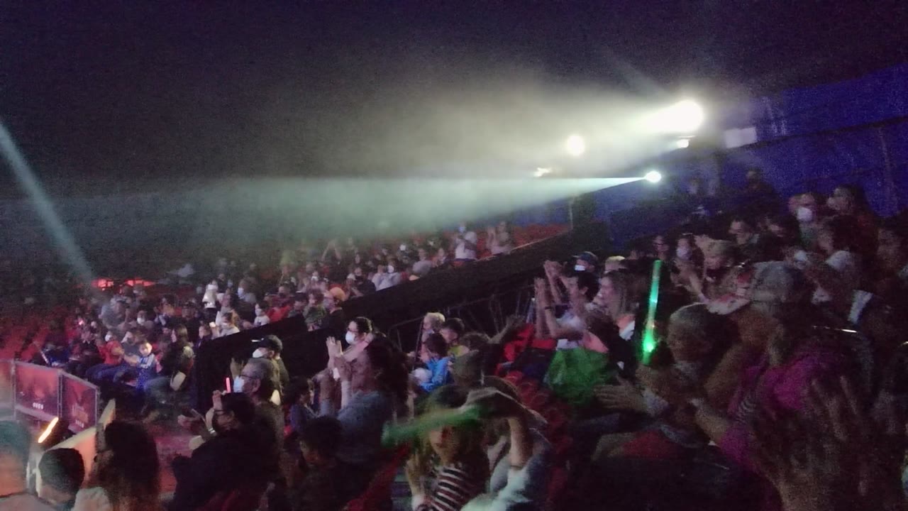 Wide stage-side view of a nighttime concert crowd in San Jose del Cabo, Baja California Sur, with clapping spectators, haze, and white spotlights sweeping over the audience.