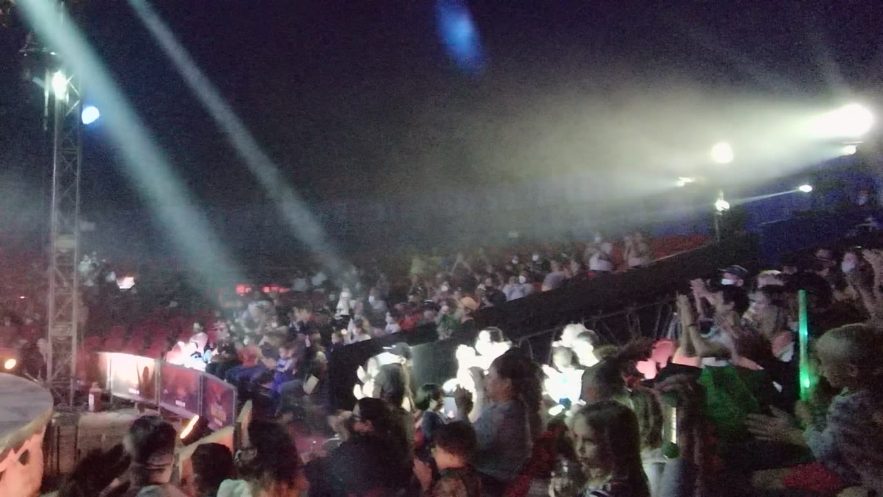 A nighttime crowd in San Jose del Cabo, Baja California Sur, watches from packed seating as bright white light washes the right side of the audience and haze hangs above them.