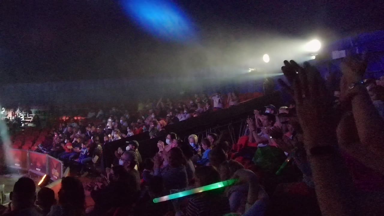 Audience members clap inside a dark San Jose Del Cabo concert venue while green glow sticks and raised hands appear in the foreground under blue and white stage light.
