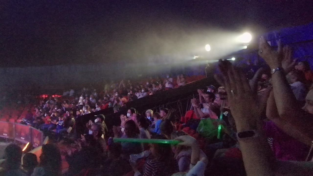 Foreground hands clap as a concert crowd watches under bright stage beams.