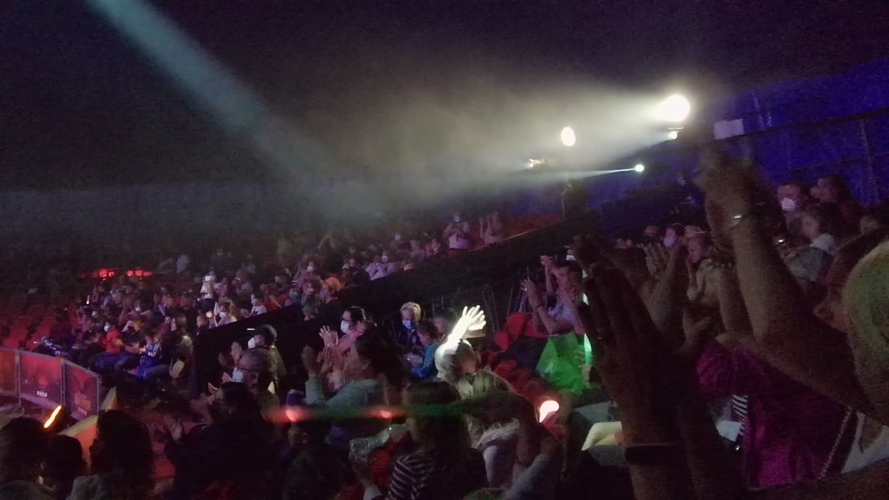 The crowd remains seated and standing in rows while hands rise near the lens and a green glow stick cuts across the lower frame in the San Jose Del Cabo venue.
