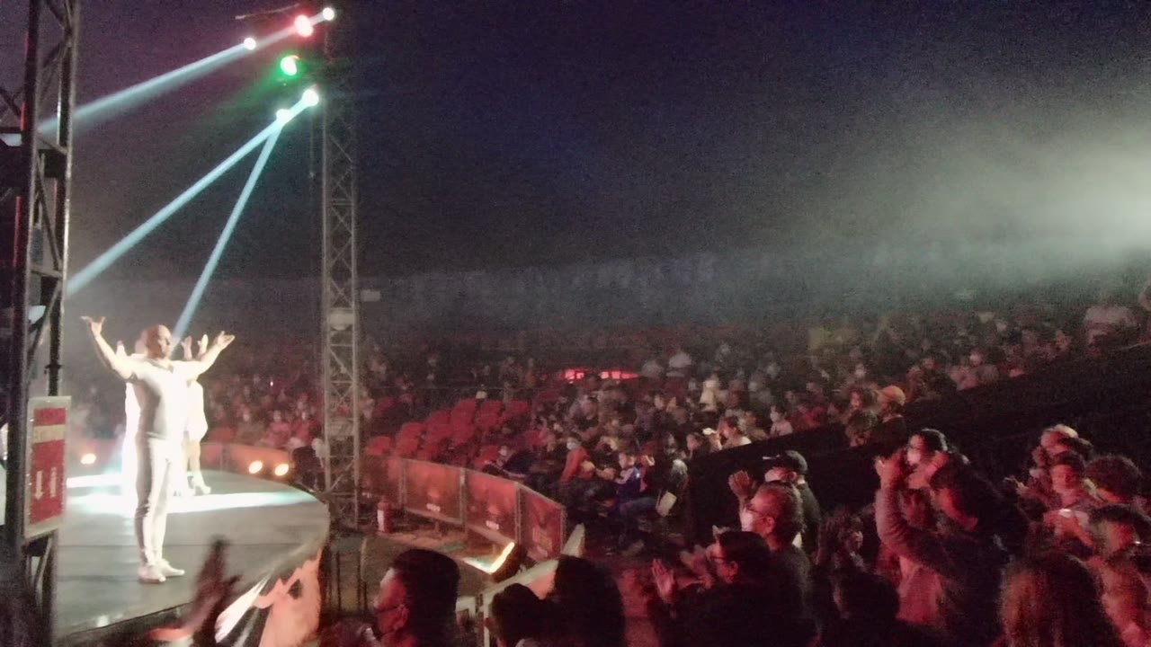 A white-clad performer stands at the edge of a curved stage in San Jose del Cabo as a large audience watches under blue-white beams and green lights.