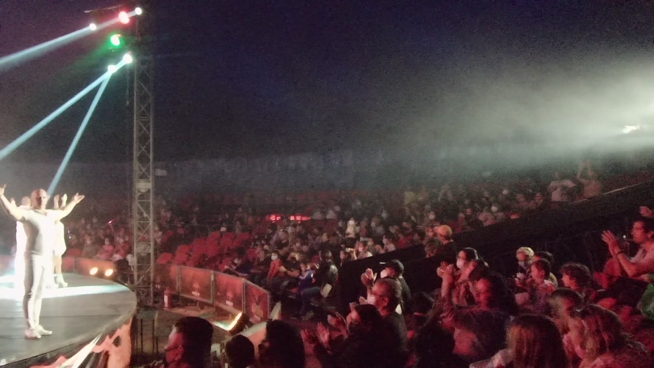 Performer on stage with arms raised toward a seated crowd and bright crossing beams.