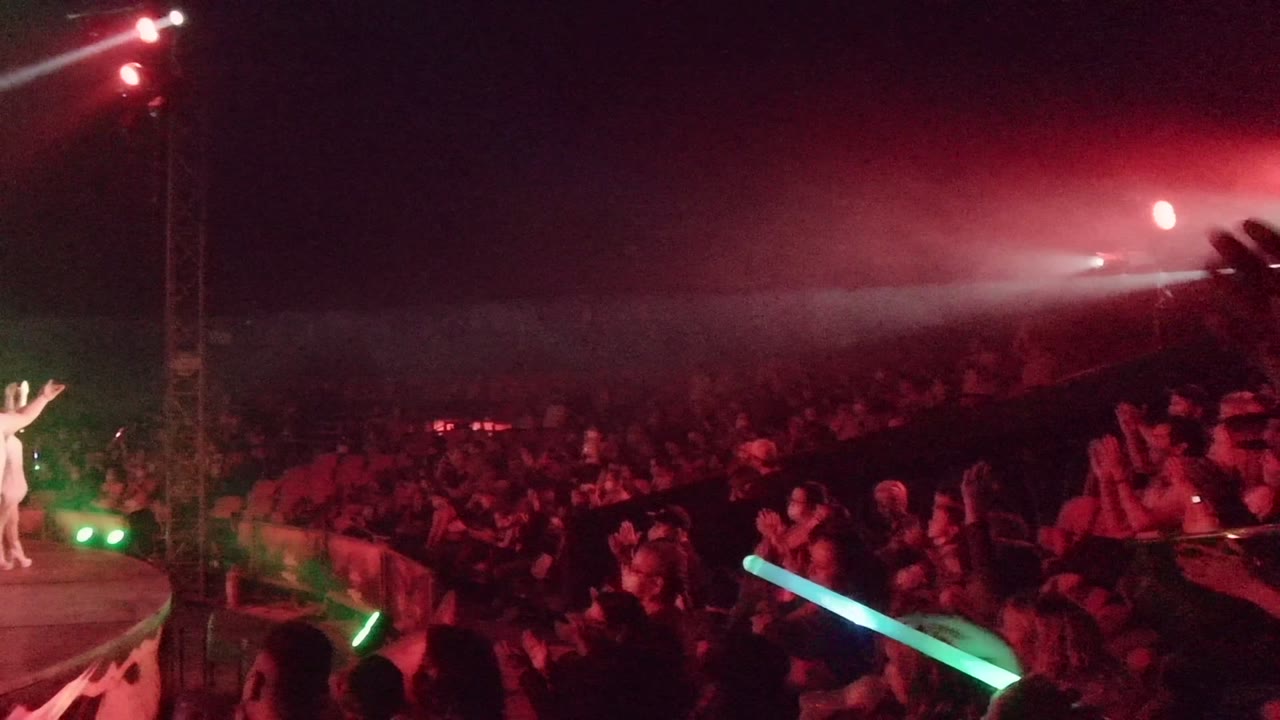 The camera view shifts toward the San Jose del Cabo audience, with clapping spectators in the foreground and the white performer farther left under red and green stage lights.