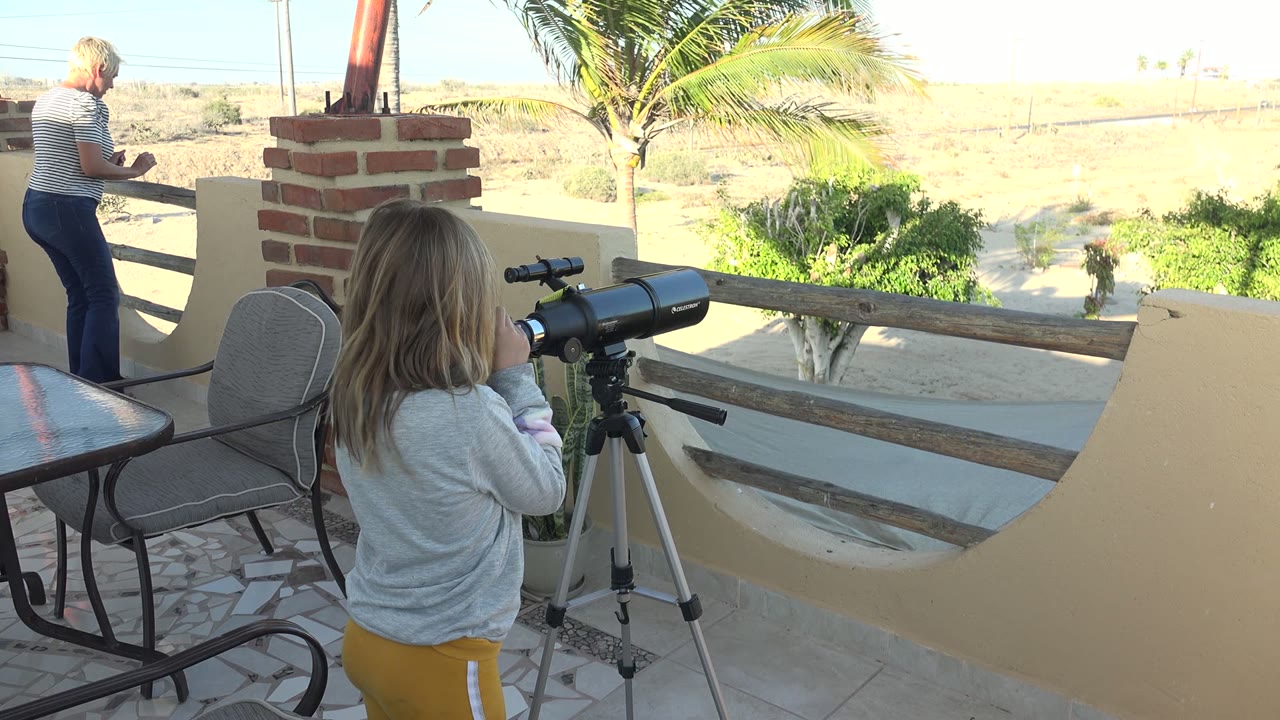 In San Carlos, Mexico, a young girl looks through a black telescope on a tripod as an adult stands at the far left near the patio railing.