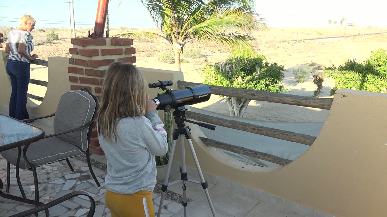Girl uses a telescope on a patio as an adult moves away at left.